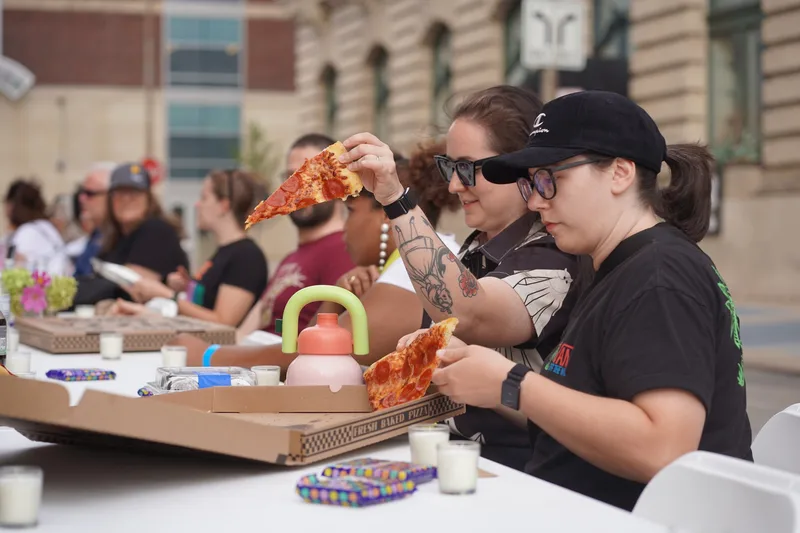 Two guests lifting slices from a large pizza box on the long table, with votive candles and candy spread around them
