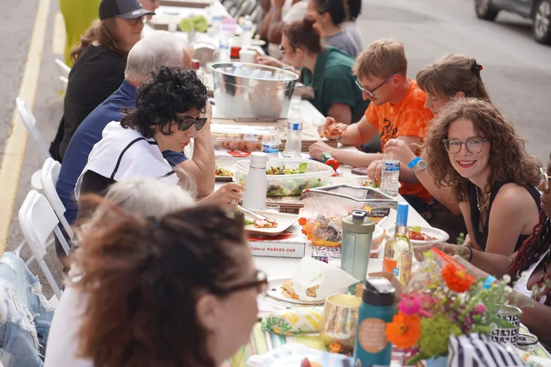 Guests eating and talking along the full length of the long table, with salads, pizza boxes, water bottles and flowers running down the middle